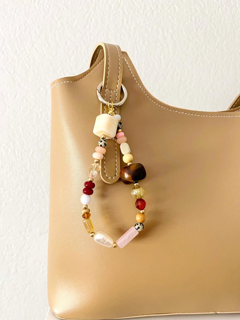 May include: A beaded keychain with a variety of colorful beads, including white, pink, red, and gold, hangs from a tan handbag. The beads are various shapes and sizes, creating a unique and eye-catching accessory.