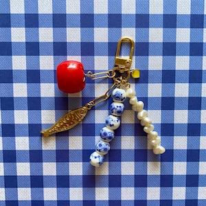 May include: A gold-toned keychain with a red cherry charm, a gold fish charm, and a strand of blue and white floral beads. A strand of white pearls and a yellow heart charm are also attached. The background is a blue and white checkered pattern.