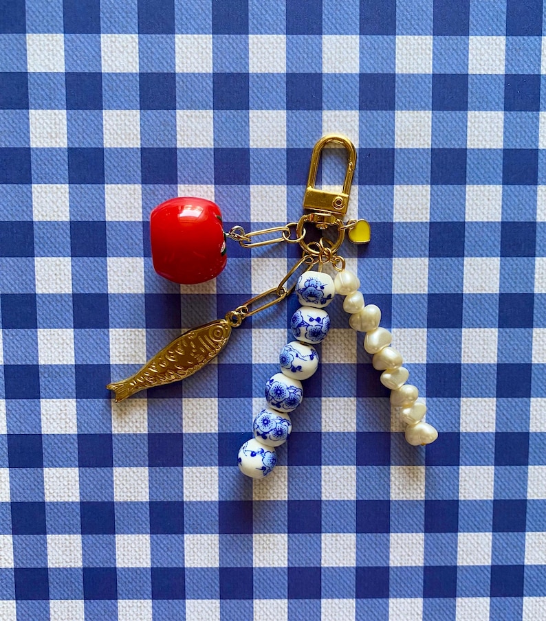 May include: A gold-toned keychain with a red cherry charm, a gold fish charm, and a strand of blue and white floral beads. A strand of white pearls and a yellow heart charm are also attached. The background is a blue and white checkered pattern.