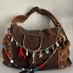 May include: A brown suede handbag with a decorative chain of colorful beads and charms, including a red coral, seashells, and a starfish. The bag has a leather handle and is adorned with gold-colored studs.