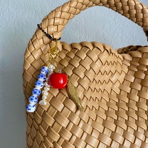 May include: Tan woven handbag with a braided handle. A gold-toned charm with a red apple, a gold fish, and blue and white beads hangs from the handle. The bag has a woven pattern.