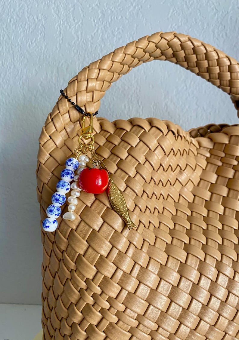 May include: Tan woven handbag with a braided handle. A gold-toned charm with a red apple, a gold fish, and blue and white beads hangs from the handle. The bag has a woven pattern.