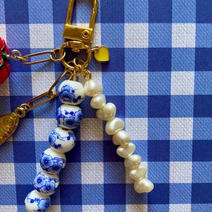 May include: A gold-colored keychain with a clasp, a small yellow heart charm, and a chain holding a strand of white pearls and blue and white ceramic beads. The beads have a floral design. The background is a blue and white checkered pattern.