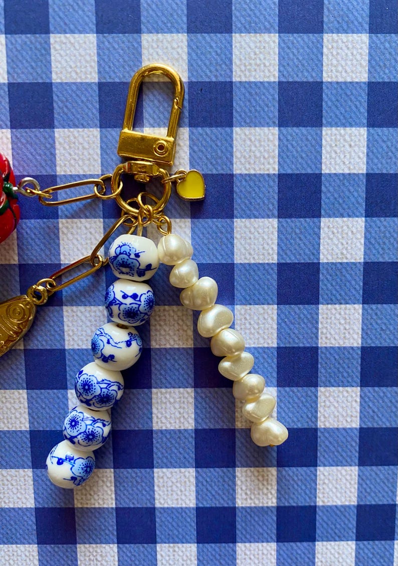 May include: A gold-colored keychain with a clasp, a small yellow heart charm, and a chain holding a strand of white pearls and blue and white ceramic beads. The beads have a floral design. The background is a blue and white checkered pattern.
