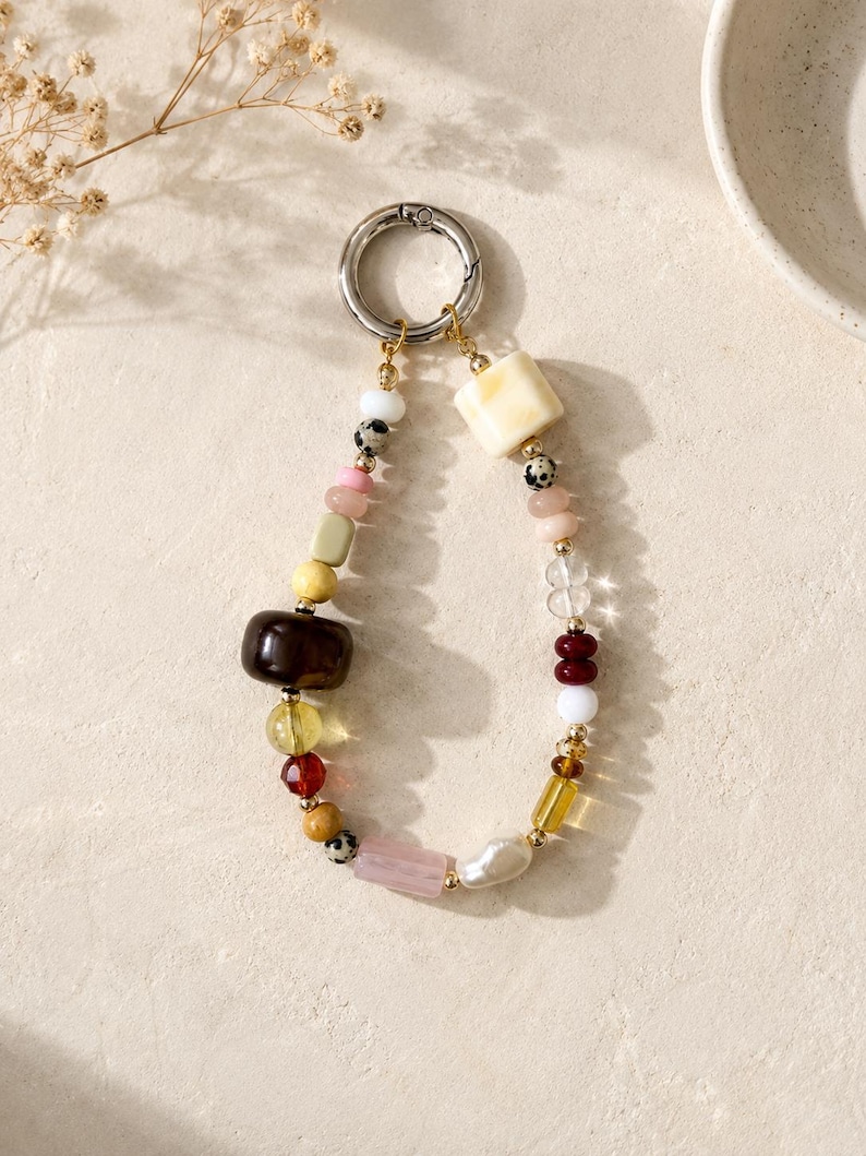 May include: A beaded keychain with a silver clasp. The beads are a mix of shapes and colors, including white, pink, yellow, brown, and clear. Some beads have a speckled pattern. The keychain is likely used for keys or as a decorative accessory.
