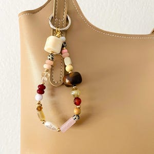May include: A beige leather handbag with a decorative beaded charm. The charm features a variety of colorful beads, including cream, pink, brown, and gold, attached to a silver ring. The bag has a curved top and a handle.