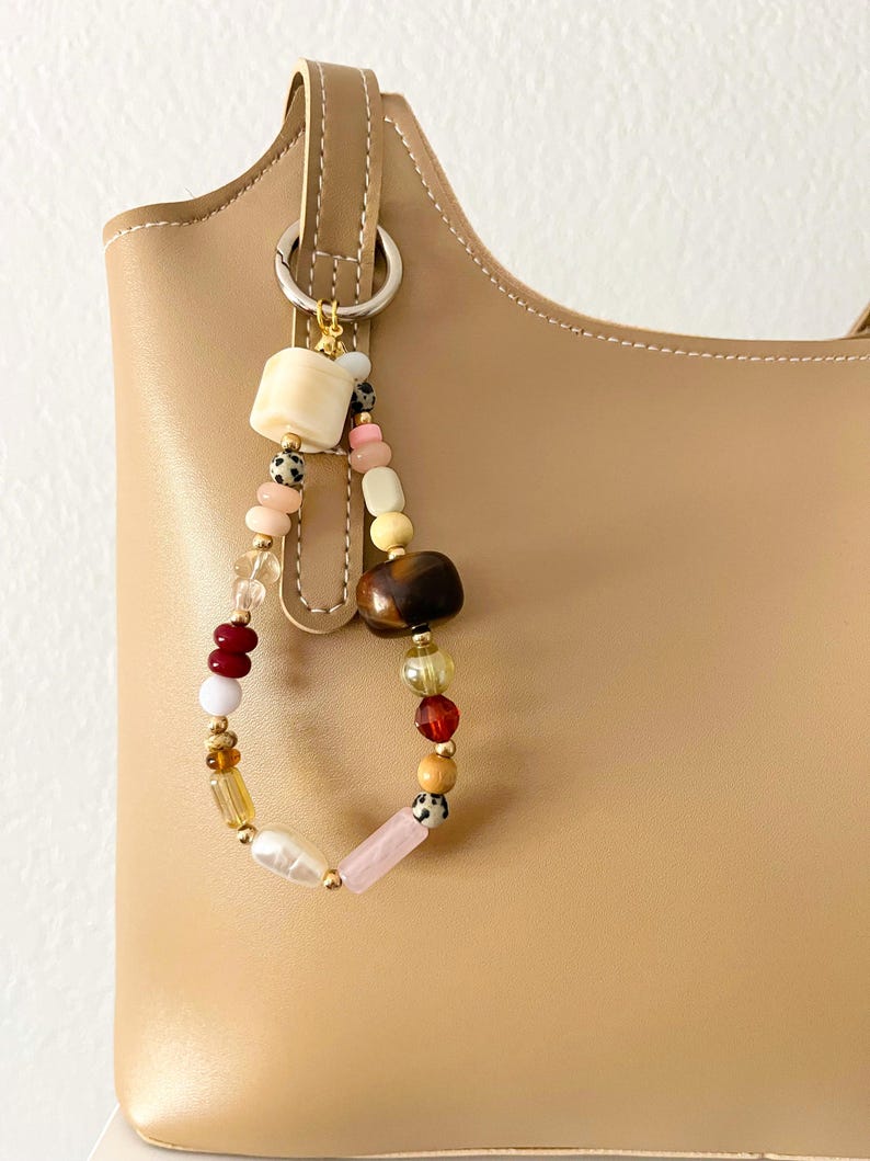 May include: A beige leather handbag with a decorative beaded charm. The charm features a variety of colorful beads, including cream, pink, brown, and gold, attached to a silver ring. The bag has a curved top and a handle.