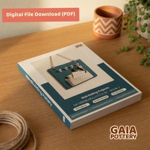 May include: A book titled "Handbuilding Pottery" with a teal cover featuring a pottery project. The book includes the text "Slab Rolling Projects" and "Air Dry Clay." A small terracotta pot and a plant are in the background.