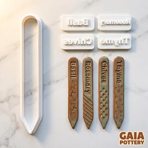 May include: Ceramic garden markers labelled Basil, Rosemary, Chives, and Thyme. The brown and green markers have embossed lettering and patterns. White molds for creating the markers are also visible.