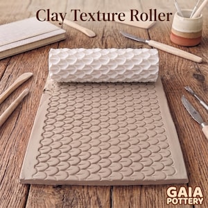 May include: A white cylindrical clay texture roller with a repeating scale pattern, positioned on a flat slab of light brown clay that displays the same embossed scale design. The text Clay Texture Roller and GAIA POTTERY are visible.