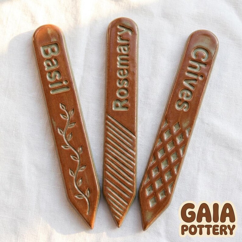 Macro close-up of three handmade Gaia Pottery ceramic herb markers for Basil, Rosemary and Chives showing rich terracotta glaze with celadon green in carved recesses and decorative textures
