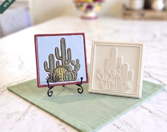 Puede incluir: Dos azulejos cuadrados de 12cm x 12cm, uno con fondo azul claro y cactus verdes y amarillos, el otro una versión en relieve blanco sólido. El azulejo de color descansa sobre un soporte de metal negro, mientras que el blanco está plano.