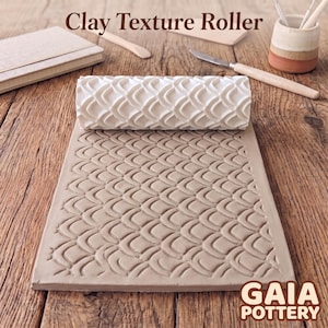 May include: A white cylindrical clay texture roller with a repeating scale pattern, positioned above a rectangular slab of light brown clay featuring the same embossed scale design. The text Clay Texture Roller and GAIA POTTERY are visible.