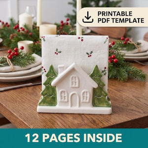 Può includere: Un portatovaglioli in ceramica bianca a forma di casa affiancata da due pini verdi. Il supporto contiene un tovagliolo di carta bianco stampato con un motivo di piccole bacche di agrifoglio rosse e foglie verdi. Il testo PRINTABLE PDF TEMPLATE è visibile.