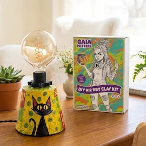 May include: A decorative lamp featuring a black cat design on a yellow base, with a large Edison bulb. A DIY air dry clay kit with a colourful illustration is placed nearby. The lamp is on a wooden table.
