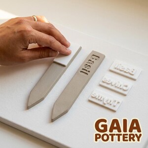 Process Shot #2: Stamping - Hand pressing Basil name stamp tile into freshly cut raw clay herb marker with Rosemary and Chives markers already stamped nearby — Gaia Pottery herb marker making kit in use