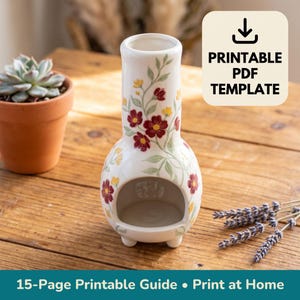 Op de afbeelding: Een witte keramische waxwarmer met een bloemenpatroon van rode en gele bloemen en groene ranken. De warmer heeft een cilindrische bovenkant, een ronde basis met een grote opening aan de voorzijde en kleine voetjes. Tekst: PRINTABLE PDF TEMPLATE.