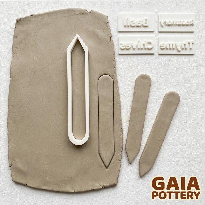 Process Shot #1: Rolling & Cutting - Hands pressing white clay herb marker cutter into grey clay slab with Basil, Rosemary, Chives and Thyme name stamps arranged nearby — Gaia Pottery herb marker making kit in action