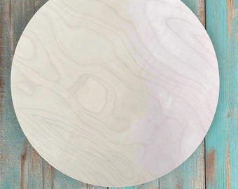May include: A circular, flat wooden board with a smooth, light-colored surface. The wood features a natural, subtle grain pattern with swirling lines in shades of cream and pale beige, showcasing the organic texture of the material.