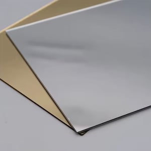 Può includere: Due fogli rettangolari di materiale riflettente, uno argentato e uno dorato, sono sovrapposti ad angolo. Il foglio argentato è sopra, oscurando parzialmente il foglio dorato. Le superfici sono lisce e lucide, riflettendo la luce.