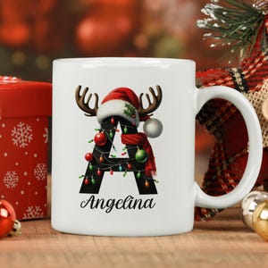 May include: White ceramic mug with a black letter A decorated with a red Santa hat, reindeer antlers, and Christmas lights. The name "Angelina" is written below the letter.