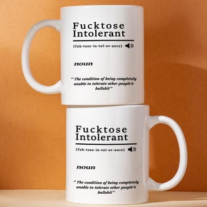 May include: Two white ceramic mugs stacked on top of each other. Each mug has a large handle and features the text "Fucktose Intolerant" with a phonetic pronunciation and a definition. The mugs are on a brown background.