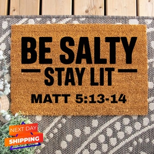 May include: A brown coir doormat with the text "BE SALTY - STAY LIT - MATT 5:13-14" in black lettering.