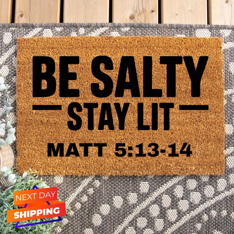 May include: A brown coir doormat with the text "BE SALTY - STAY LIT - MATT 5:13-14" in black lettering.