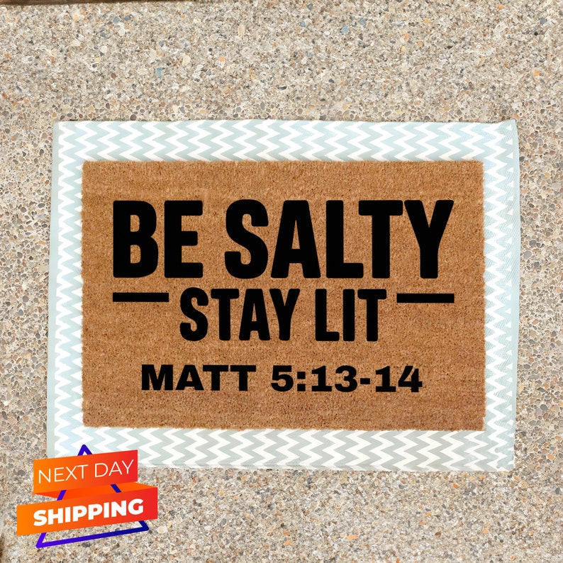 May include: A brown coir doormat with the text "BE SALTY - STAY LIT - MATT 5:13-14" in black lettering. The doormat has a light gray and white chevron border.