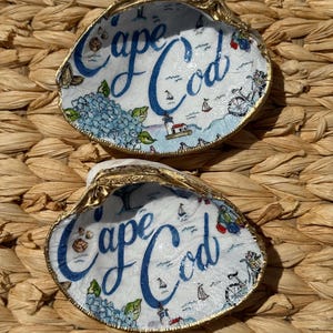 May include: Two decorative oyster shells with gold edging. Each shell features a coastal design, including the words "Cape Cod" in blue script, sailboats, a lighthouse, and blue hydrangea flowers.