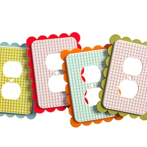 Puede incluir: Cuatro cubiertas de enchufes de colores. Cada cubierta tiene un borde festoneado y un patrón de cuadrícula. Los colores incluyen amarillo, rojo, naranja y verde. Las cubiertas tienen dos aberturas circulares para los enchufes.
