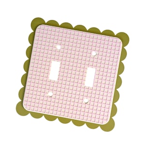 May include: A double light switch wall plate featuring a light pink grid pattern on a white background, framed by an olive green scalloped border. The rectangular plate includes two vertical toggle switch openings and four screw holes for mounting.