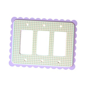 May include: A triple light switch wall plate featuring a light green grid pattern on a beige background. The rectangular plate has a scalloped lavender border and includes three vertical rectangular openings for switches and six small screw holes.