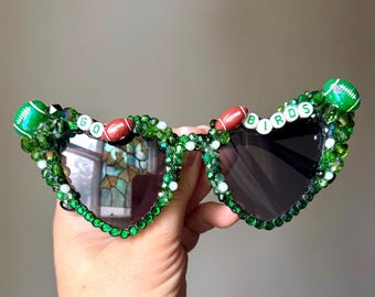 May include: Heart-shaped sunglasses with dark lenses, embellished with green beads and football accents. The text "GO BIRDS" is spelled out across the top of the frame. The sunglasses are held up by a hand.
