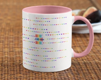 Puede incluir: Taza de cerámica blanca con borde y asa de color rosa. La taza tiene un diseño de bandera arcoíris con las palabras "Just Love" en ella. El fondo está cubierto con un patrón de pequeños corazones de colores.