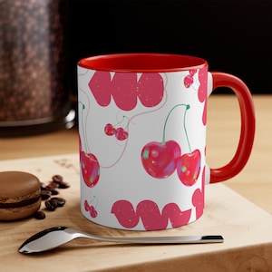 May include: A red and white ceramic mug with a cherry pattern. The mug has a red rim and handle. The cherries are pink and white with a glossy finish.