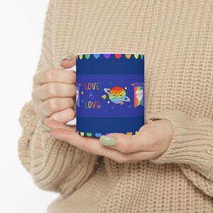 May include: A white ceramic mug with a blue background and a rainbow planet design. The mug has the text "LOVE IS LOVE" in rainbow letters and a rainbow heart border.