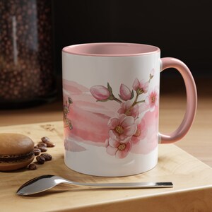 May include: A pink and white ceramic mug with a watercolor design of pink flowers and leaves. The mug has a pink rim and handle.