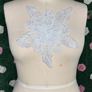 May include: A white, sequined floral applique is displayed on a dress form. The applique is in the shape of a large flower with detailed petals. The dress form has the words "COLLAPSIBLE SHOULDER" and the number "18" printed on it.