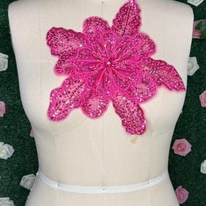 Könnte beinhalten: Eine leuchtend pinkfarbene Applikation in Form einer Blume mit Pailletten wird auf einer Schaufensterpuppe präsentiert. Die Blume ist groß und detailliert, mit mehreren Blütenblättern und einem funkelnden Finish. Die Schaufensterpuppe ist beige, mit den Worten "COLLAPSIBLE SHOULDER" und der Zahl "18" darauf.