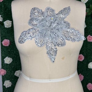 May include: A silver sequined floral applique is displayed on a dress form. The applique is in the shape of a large flower with multiple petals. The dress form is beige and has the words "COLLAPSIBLE SHOULDER" and the number "18" printed on it.
