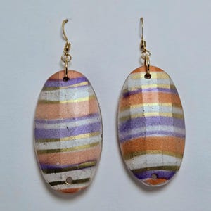 May include: A pair of oval-shaped earrings with a colorful striped design. The earrings feature horizontal stripes in shades of peach, purple, gold, and white. Gold-colored hooks complete the jewelry.