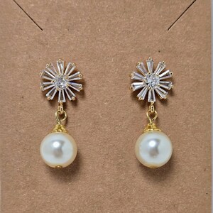May include: A pair of gold-coloured earrings. Each earring has a sparkling, starburst-shaped top with a central clear stone and a dangling, round, off-white pearl. The earrings are displayed on a brown card.