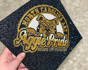May include: A navy blue graduation cap adorned with tiny, shimmering rhinestones. The cap displays a gold and white emblem with the words "North Carolina A&T State University" and "Aggie Pride" around a bulldog.