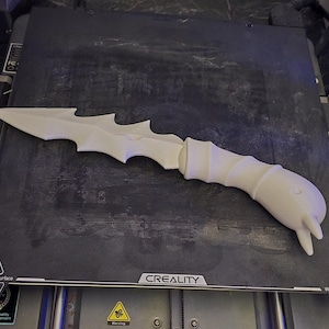 May include: A white, 3D-printed fantasy dagger with a stylized blade and handle. The dagger is resting on a black surface, likely a 3D printer bed. The handle has a textured, spiraling design.