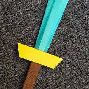 May include: A teal and yellow cardboard sword with a brown handle. The sword is lying on a gray carpet.