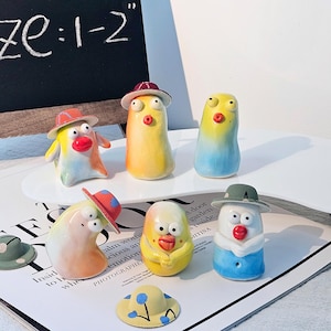 Könnte beinhalten: Eine Sammlung skurriler Keramikfiguren, jede mit einem einzigartigen Design und einem Miniaturhut. Die Figuren sind in verschiedenen Farben gehalten, darunter Gelb, Blau und Orange. Das Schild sagt "Size: 1-2".