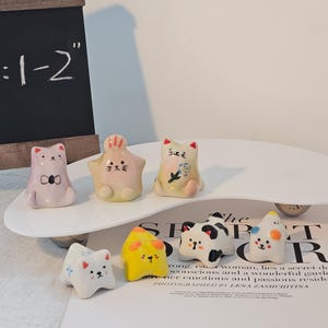 May include: A collection of small, ceramic figurines in the shape of cats and other animals. The figurines are in various colors, including pink, yellow, white, and black. The size is 1-2 inches. They are arranged on a white, curved platform.