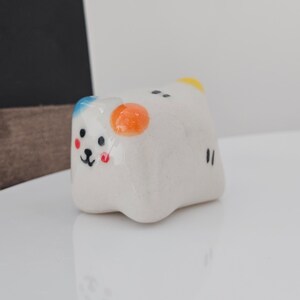 May include: A small, white ceramic figurine shaped like a bear with a smiling face. The bear has blue, orange, and yellow accents, with black eyes and a black mouth. The figurine is on a white surface.