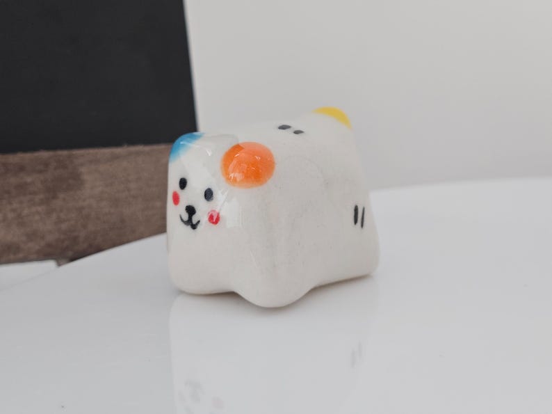 May include: A small, white ceramic figurine shaped like a bear with a smiling face. The bear has blue, orange, and yellow accents, with black eyes and a black mouth. The figurine is on a white surface.